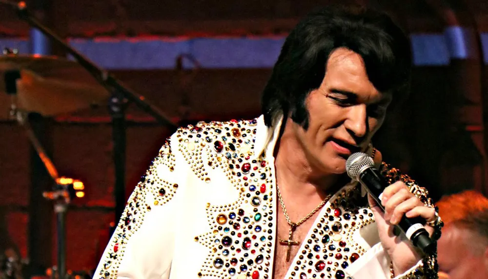 A person resembling Elvis Presley is singing into a microphone while wearing a white jewel-encrusted jumpsuit