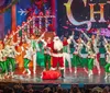 Performers in vibrant Christmas-themed costumes enthusiastically act on stage with a small inset showing others singing in a festive setting