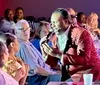 A performer in a red patterned jacket sings into a microphone while engaging with an audience sitting at tables