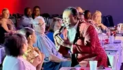 A performer in a red patterned jacket sings into a microphone while engaging with an audience sitting at tables.