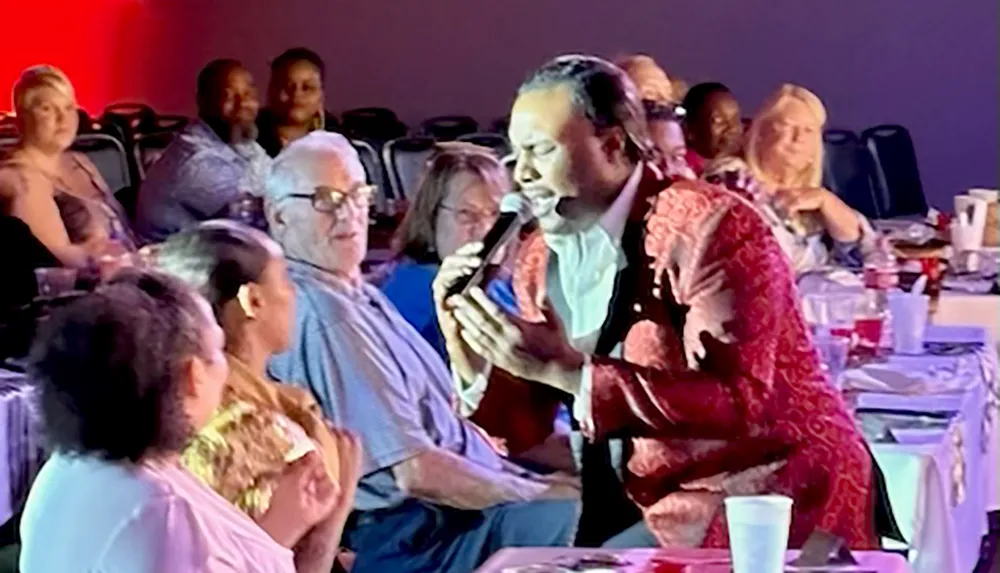 A performer in a red patterned jacket sings into a microphone while engaging with an audience sitting at tables