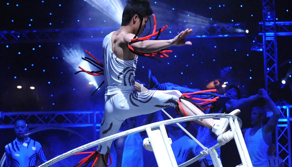 A performer is executing a dynamic airborne maneuver on a tightrope at a circus or acrobatic show surrounded by other performers in an illuminated stage environment