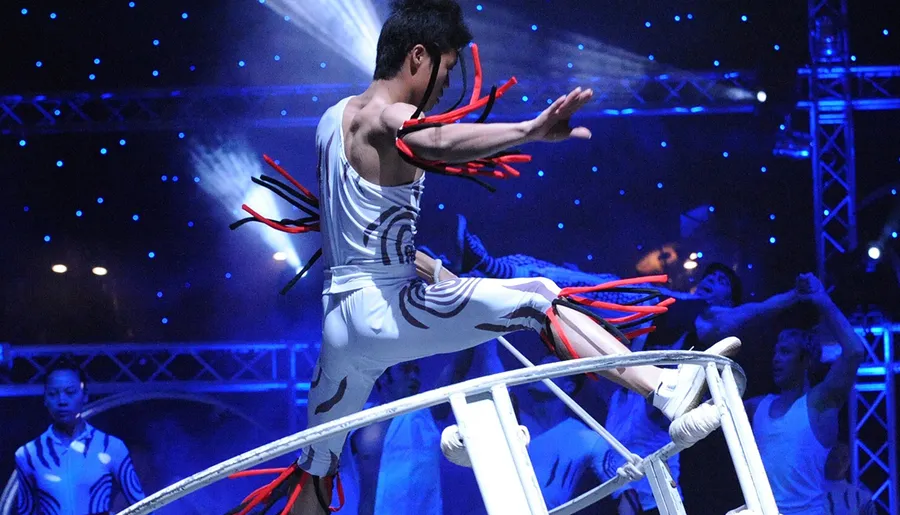 A performer is executing a dynamic, airborne maneuver on a tightrope at a circus or acrobatic show, surrounded by other performers in an illuminated stage environment.