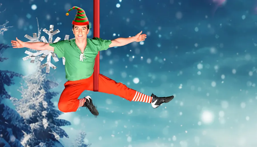 A person dressed as a festive elf is performing an acrobatic split on a vertical pole with a snowy, winter background.