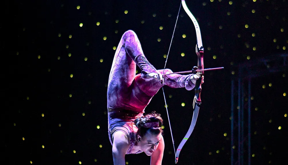 A contortionist is performing an archery trick holding a bow with her feet while in a backbend position