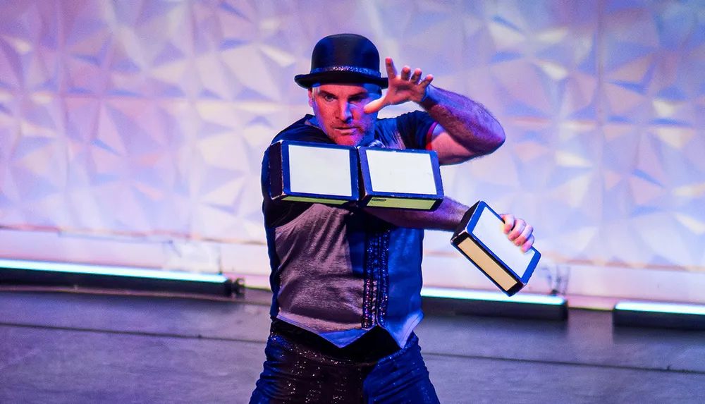 A performer wearing a hat is dramatically gesturing while holding rectangular objects in a brightly lit setting