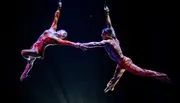 Two aerialists perform an acrobatic routine, suspended in mid-air and holding hands.