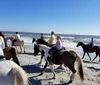 A group of people are horseback riding along a beach with a pier in the background