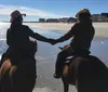 A group of people are horseback riding along a beach with a pier in the background