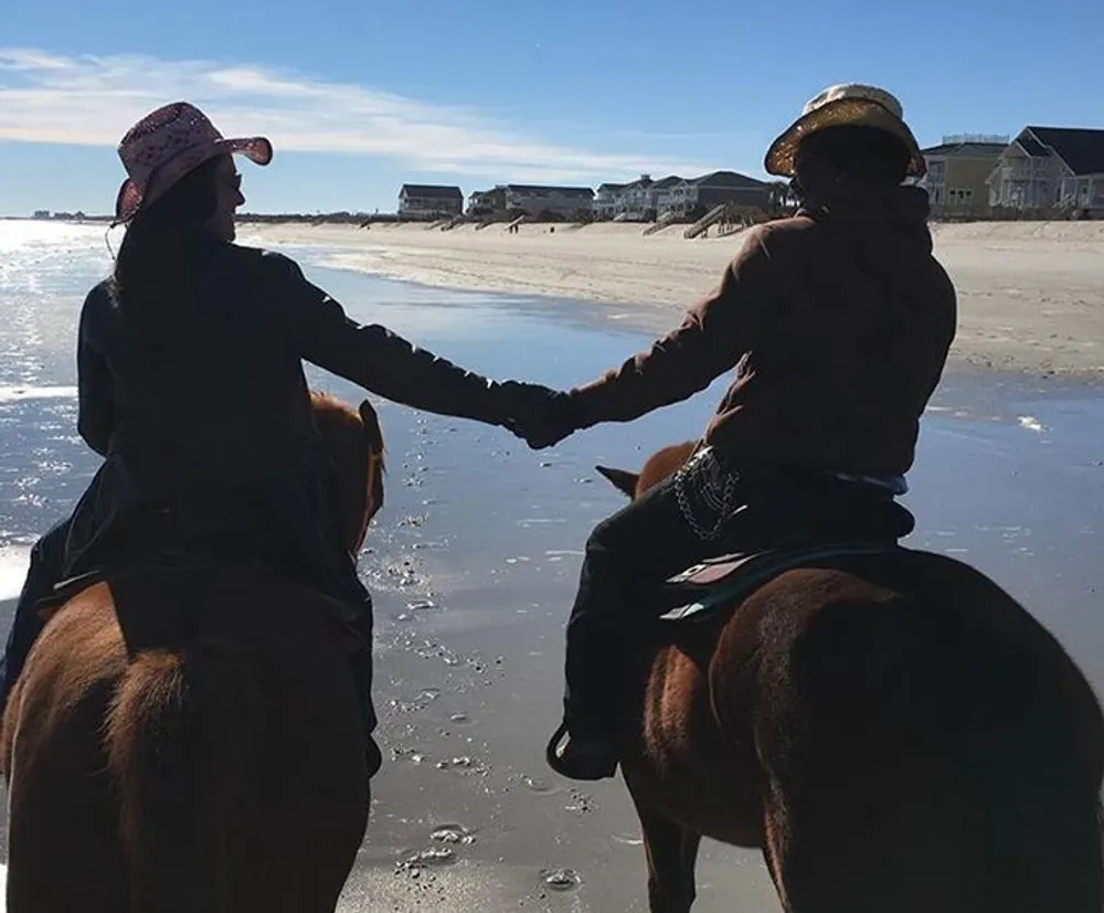 Two people are shaking hands while on horseback on a sunny beach with houses in the background