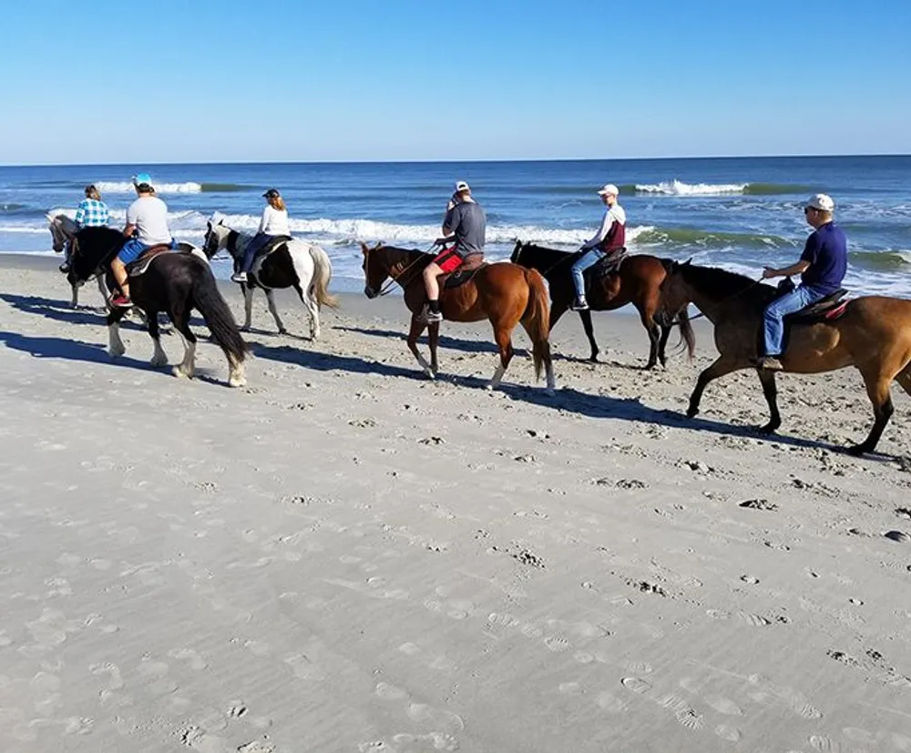 A group of people are riding horses along a sandy beach with gentle waves in the background