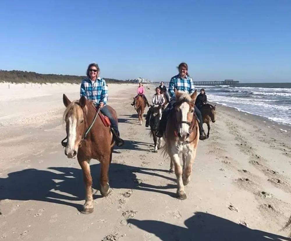 A group of people are horseback riding along a beach with a pier in the background
