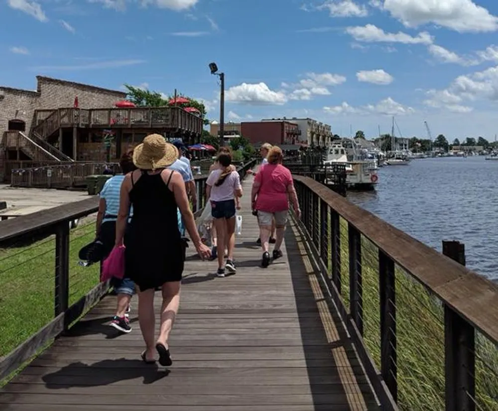 A group of people walk on a wooden boardwalk by a waterfront with boats and buildings in the background on a sunny day