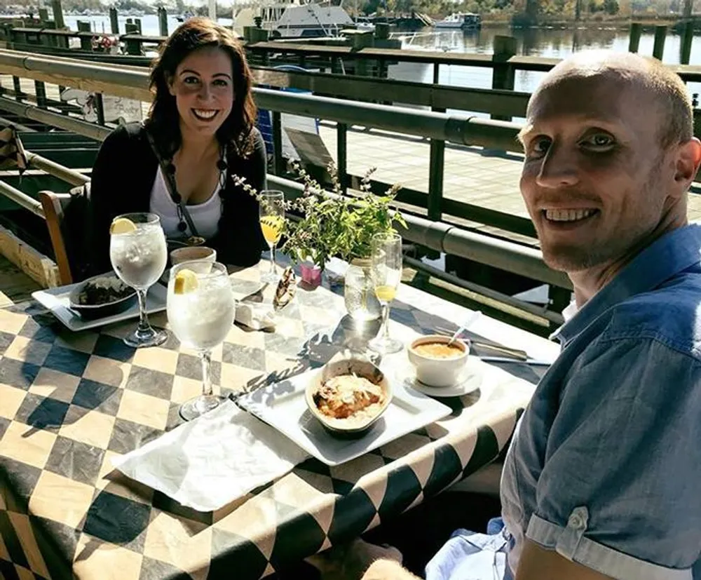 A man and a woman are smiling at the camera while enjoying a meal and drinks at an outdoor restaurant table with a water view in the background