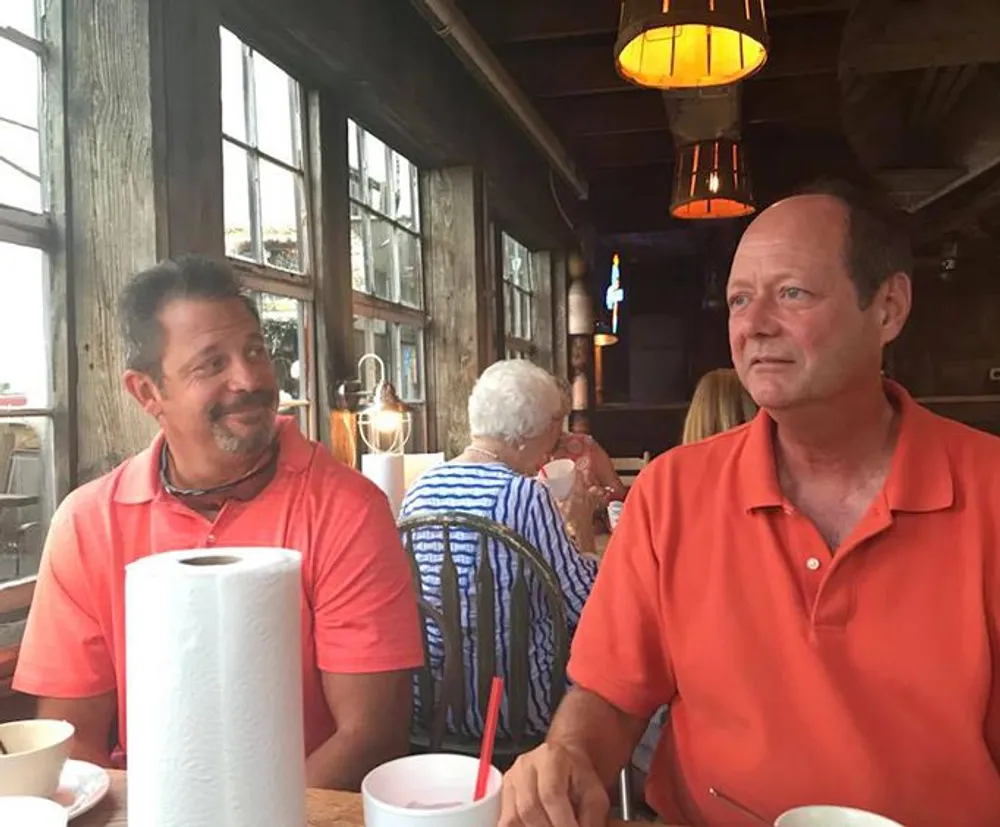 Two men are sitting at a dining table with a paper towel roll between them one looking at the camera with a neutral expression and the other smiling slightly while looking away in a rustic-style restaurant with other patrons in the background