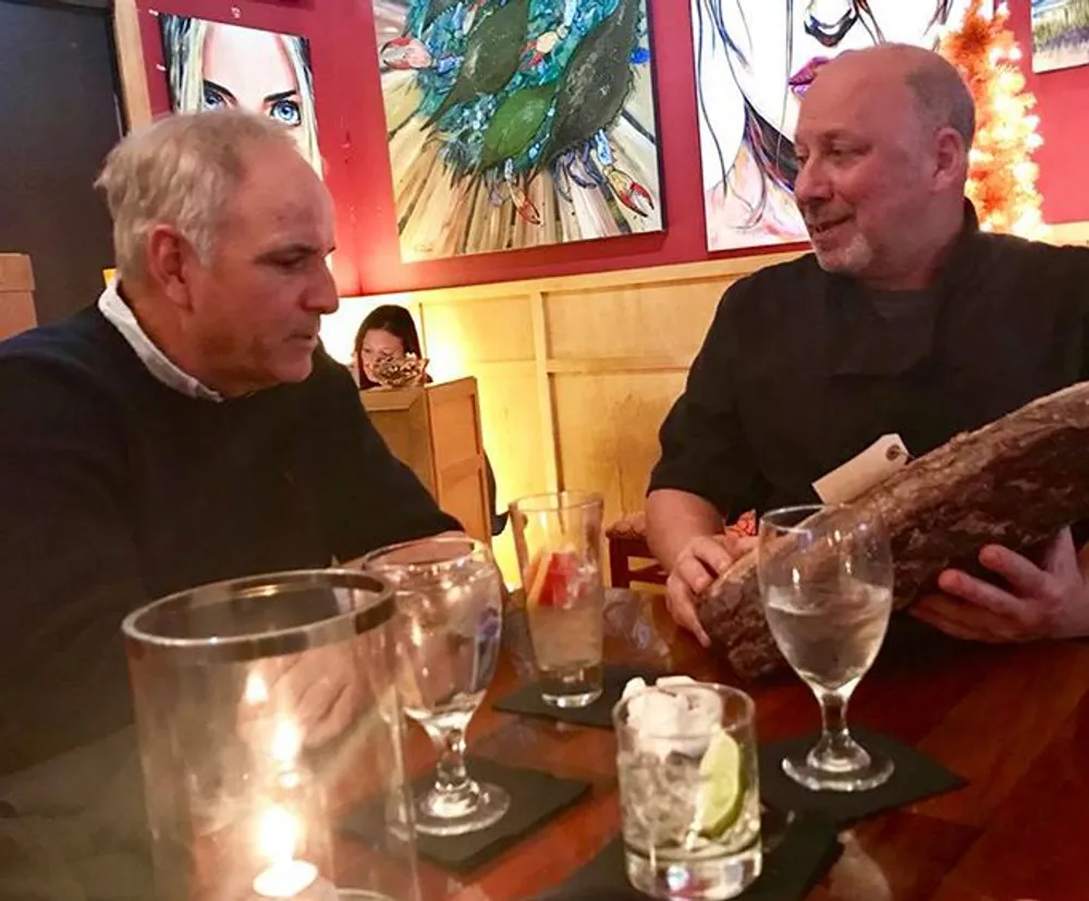Two men are engaged in conversation at a restaurant table with empty glasses and a lit candle between them and one is holding a large rustic-looking menu