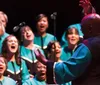 A choir passionately sings on stage as a conductor with their back to the camera leads them with expressive hand gestures