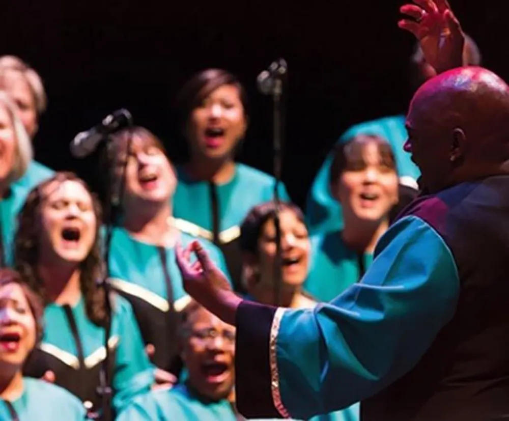 A choir passionately sings on stage as a conductor with their back to the camera leads them with expressive hand gestures