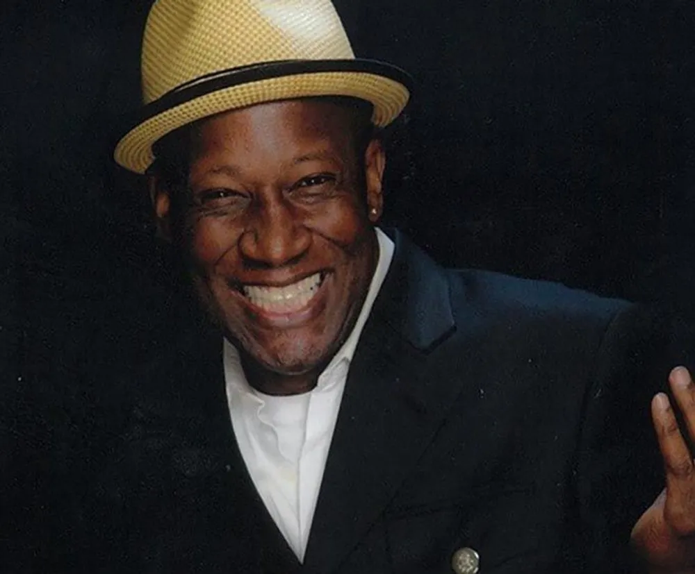 The image shows a joyful man wearing a straw hat and a dark suit smiling broadly against a black background