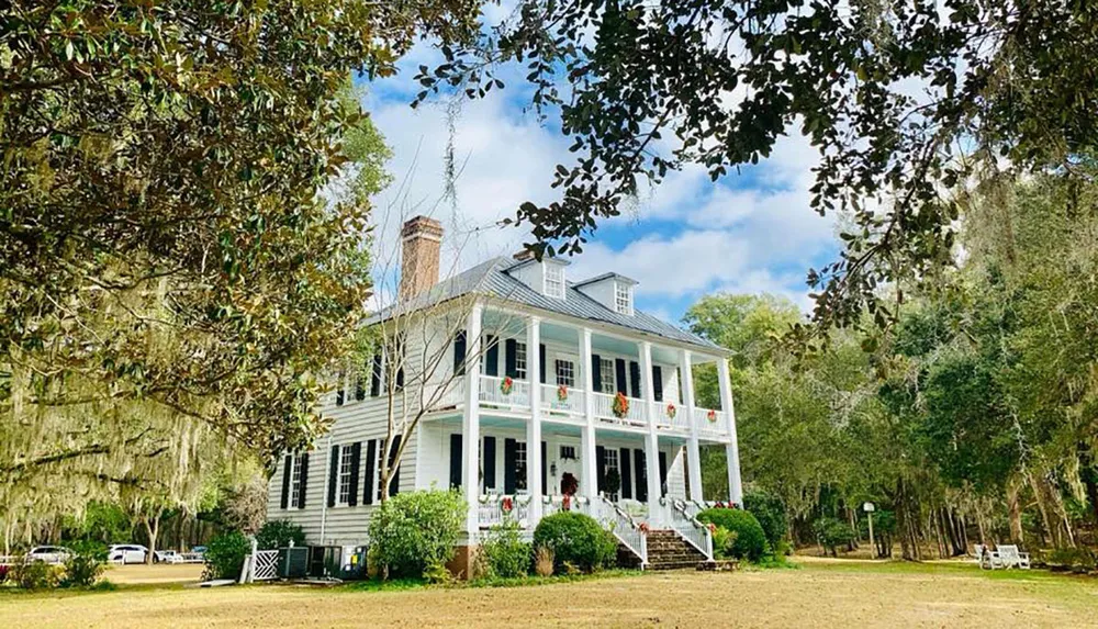The image showcases a classic two-story white house with a double porch adorned with wreaths surrounded by a serene landscape with Spanish moss-covered trees