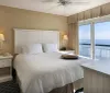 The image shows a neatly arranged hotel room with a large bed a television and a balcony that offers a picturesque view of the sea