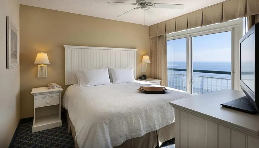 The image shows a neatly arranged hotel room with a large bed, a television, and a balcony that offers a picturesque view of the sea.