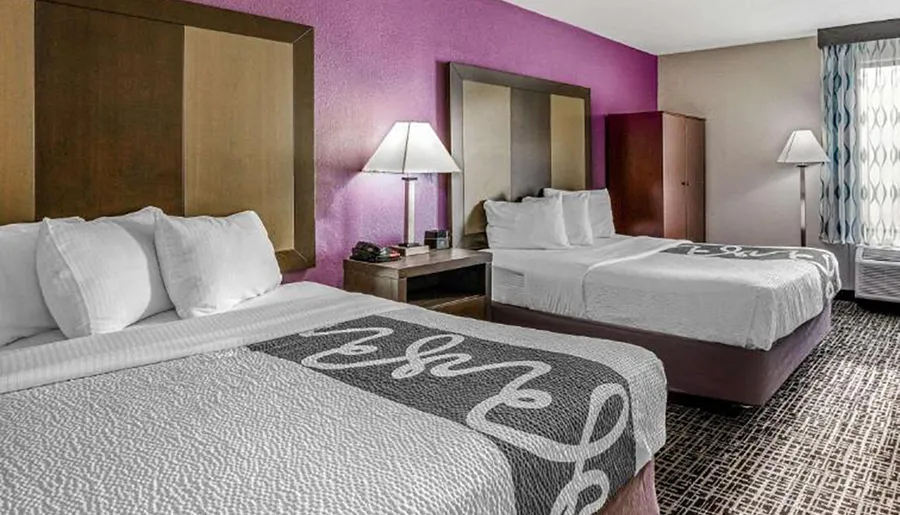 The image shows a neatly arranged hotel room with two double beds featuring white linens and a decorative throw, flanked by bedside tables with lamps, and a purple accent wall in the background.
