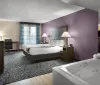 The image shows a neatly arranged hotel room with two double beds featuring white linens and a decorative throw flanked by bedside tables with lamps and a purple accent wall in the background