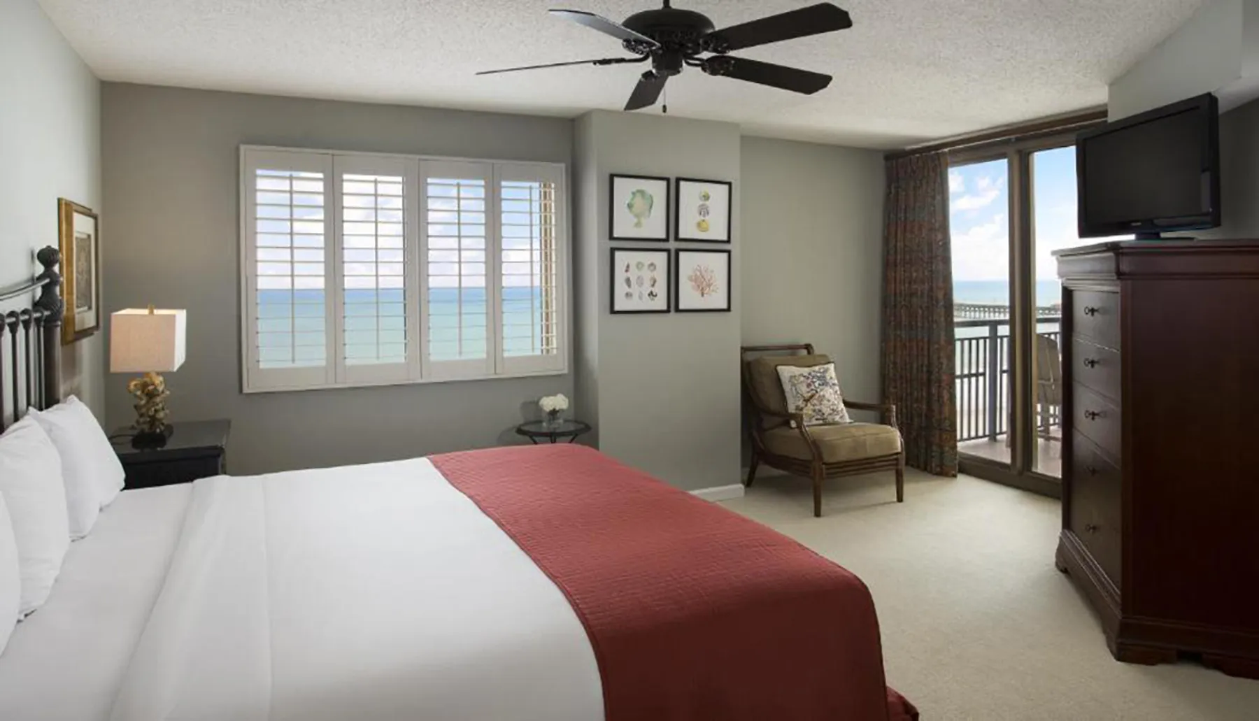 The image shows a neatly arranged bedroom with a large bed, furniture, artwork on the walls, and a balcony overlooking the ocean.