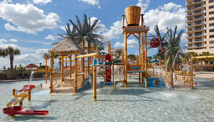 The image shows a colorful childrens water playground with various water features slides and playful structures located beside a beachfront high-rise building