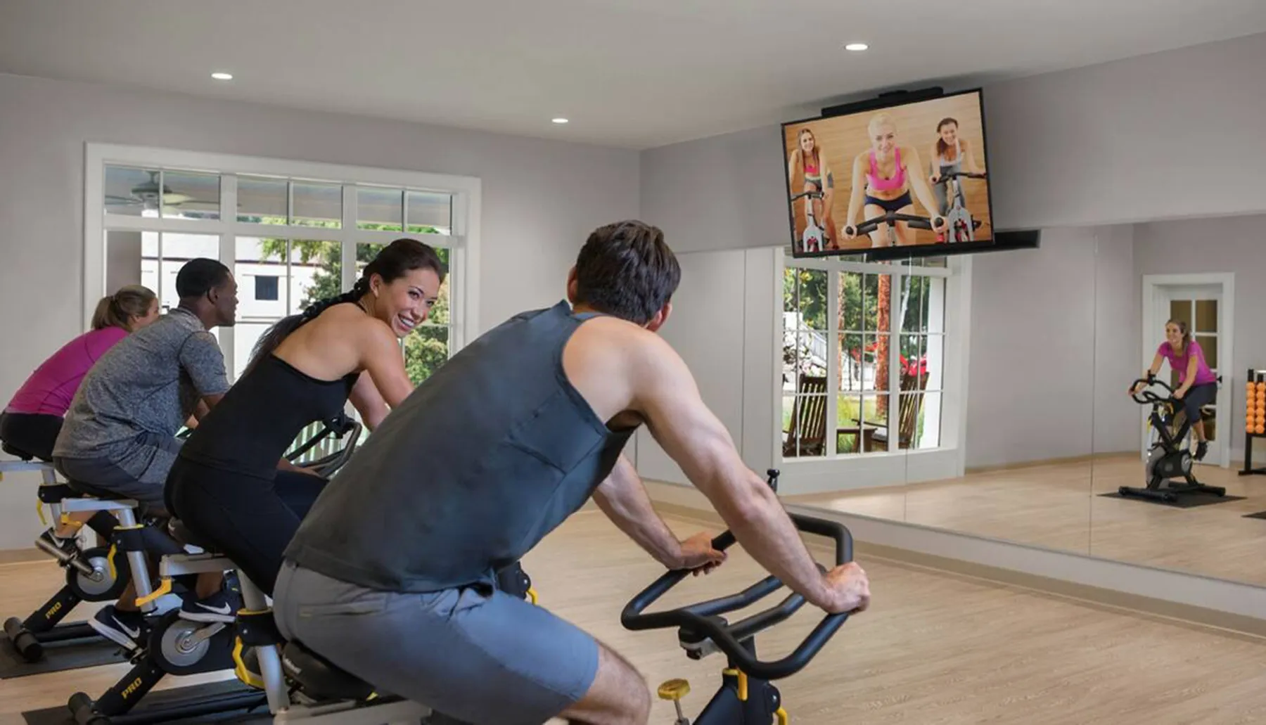 A group of people are riding stationary bikes in a fitness class while watching an instructor on a large screen in a well-lit room.