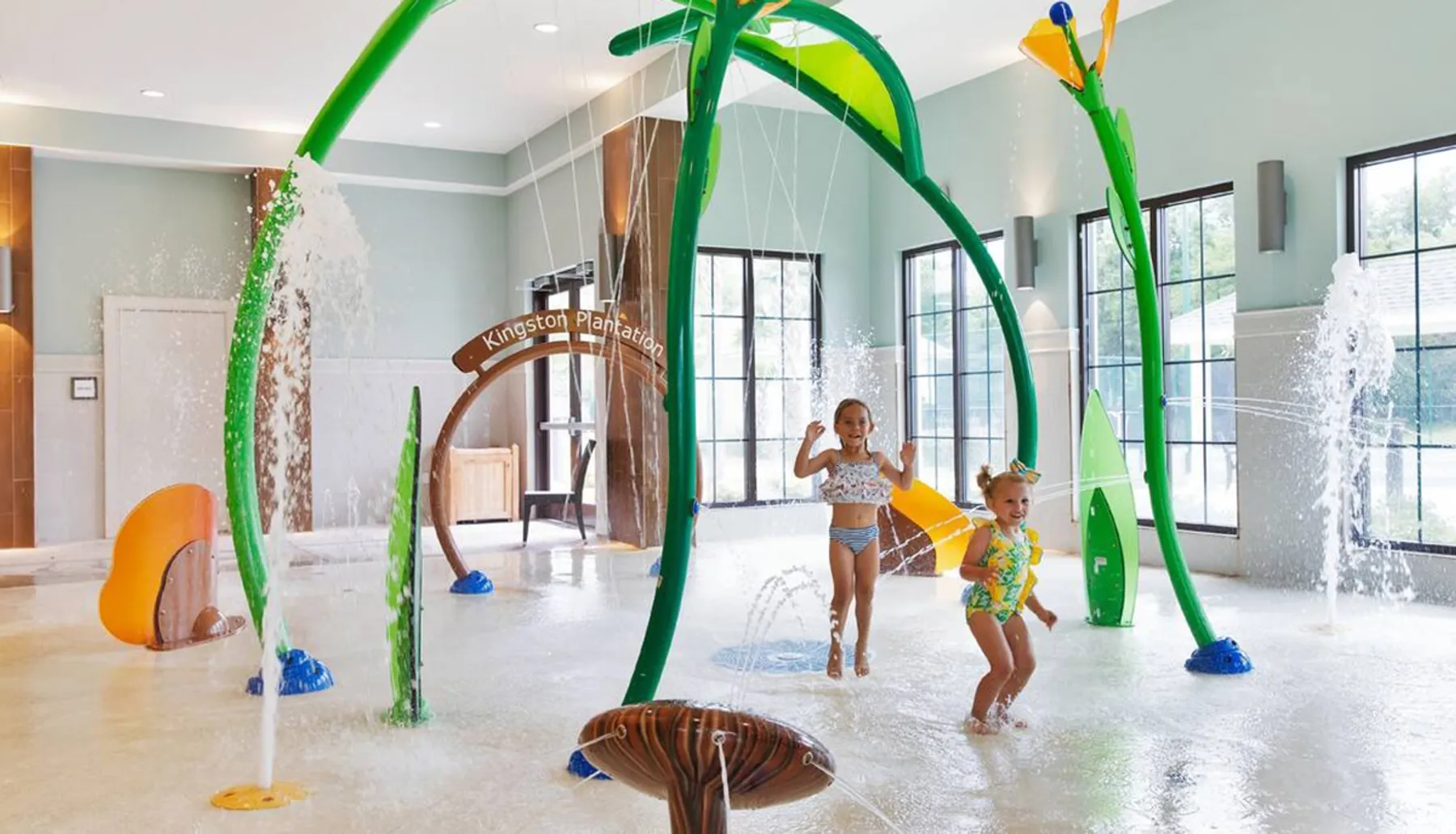 Two children play gleefully in an indoor water park with colorful water features and sprays around them.