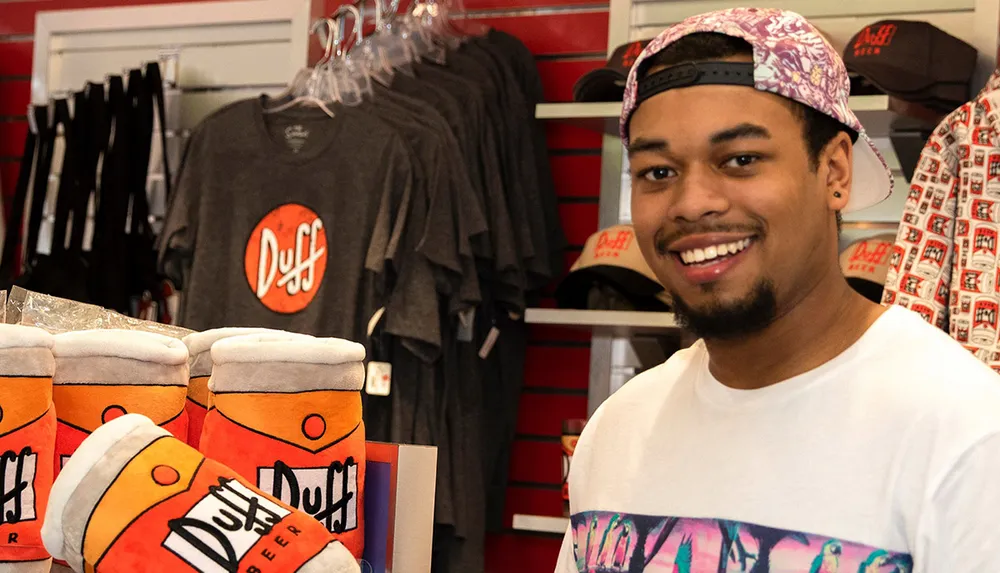 A smiling person is standing in a store filled with merchandise themed after the fictional Duff Beer from the animated series The Simpsons
