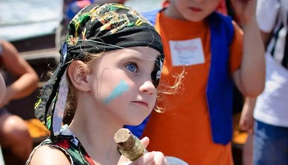 A child with face paint and a pirate bandana holds a stick and looks away thoughtfully evoking a playful pirate theme