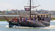 A group of people enjoy a boat ride on a miniature pirate ship flying a Jolly Roger flag.