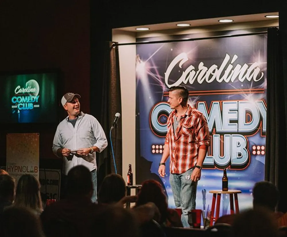 Two individuals are standing on stage with microphones at the Carolina Comedy Club while an audience watches them
