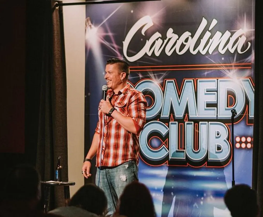 A person is performing stand-up comedy at the Carolina Comedy Club as indicated by the prominent backdrop