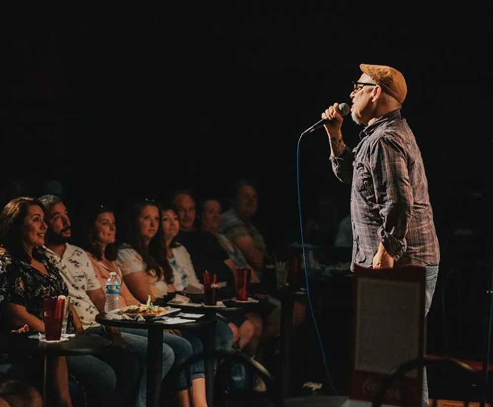 A person is performing on stage with a microphone in front of an attentive audience seated at tables