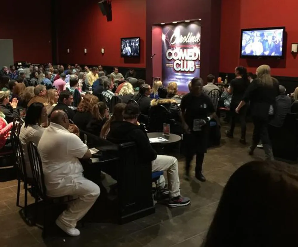 A crowded comedy club with an attentive audience facing the stage where a performance is seemingly underway with monitors displaying the act to those seated further back