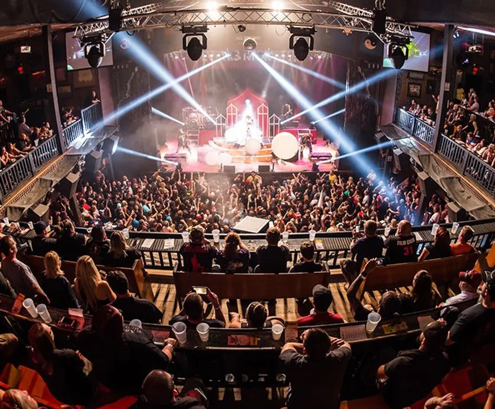 A band performs on stage at a well-lit indoor music venue with a crowd of spectators watching from the floor and balconies
