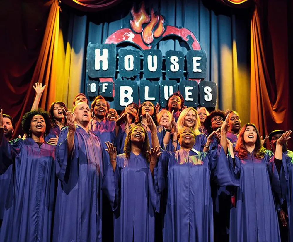 A diverse choir is joyfully singing on stage under the lit signage of the House of Blues