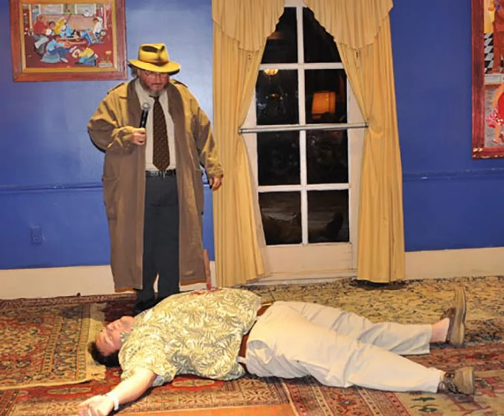A man in a trench coat and hat appears to be investigating a scene where another person is lying motionless on the floor in what seems to be a staged play or a theatrical murder mystery setting