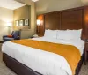 The image shows a neatly arranged hotel room with a large bed featuring white linens and an orange bed runner accompanied by modern furnishings and decor