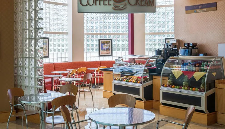 The image displays a cozy coffee shop interior with colorful chairs, glass block windows providing natural lighting, and a glass-enclosed display featuring an assortment of pastries and beverages.