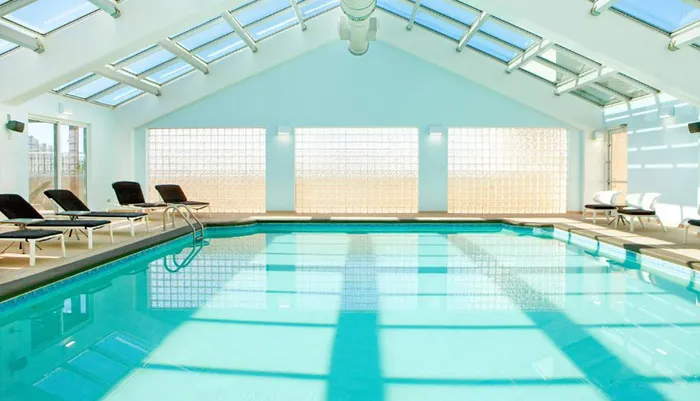 The image shows a bright and airy indoor swimming pool with a vaulted skylight ceiling several lounge chairs along the side and glass block windows filtering in natural light