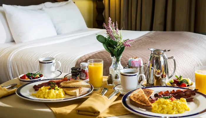 A sumptuous breakfast tray with scrambled eggs bacon toast freshly sliced fruits jam orange juice and coffee is elegantly presented on a bed with plush pillows and a warm ambiance