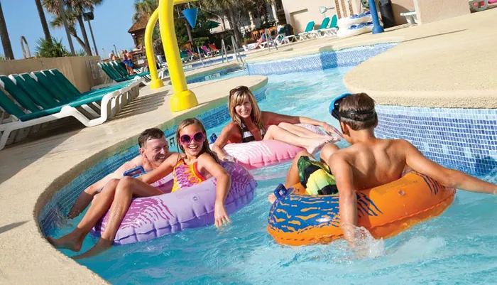 A group of people including children and adults are enjoying themselves on colorful floatation tubes in a sunny pool with a lazy river feature