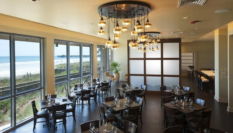 An elegant beachfront dining room with tables set for service, modern lighting fixtures, and a view of the ocean and a pier through large windows.