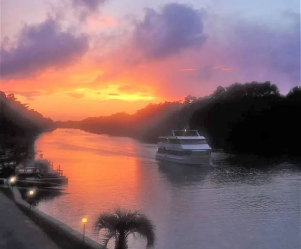 A boat cruises along a calm river under a vividly colored sunset sky