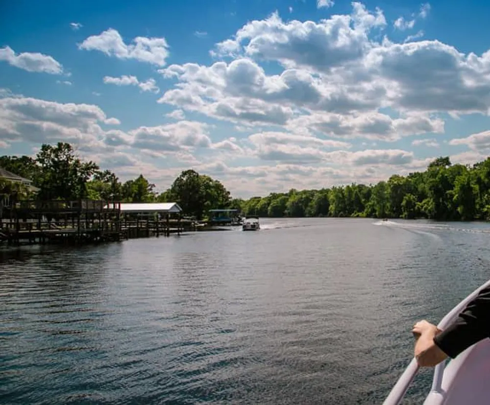 The image shows a serene river scene with a boat moving away a dock to the side and a partly cloudy blue sky overhead as seen from another boats perspective